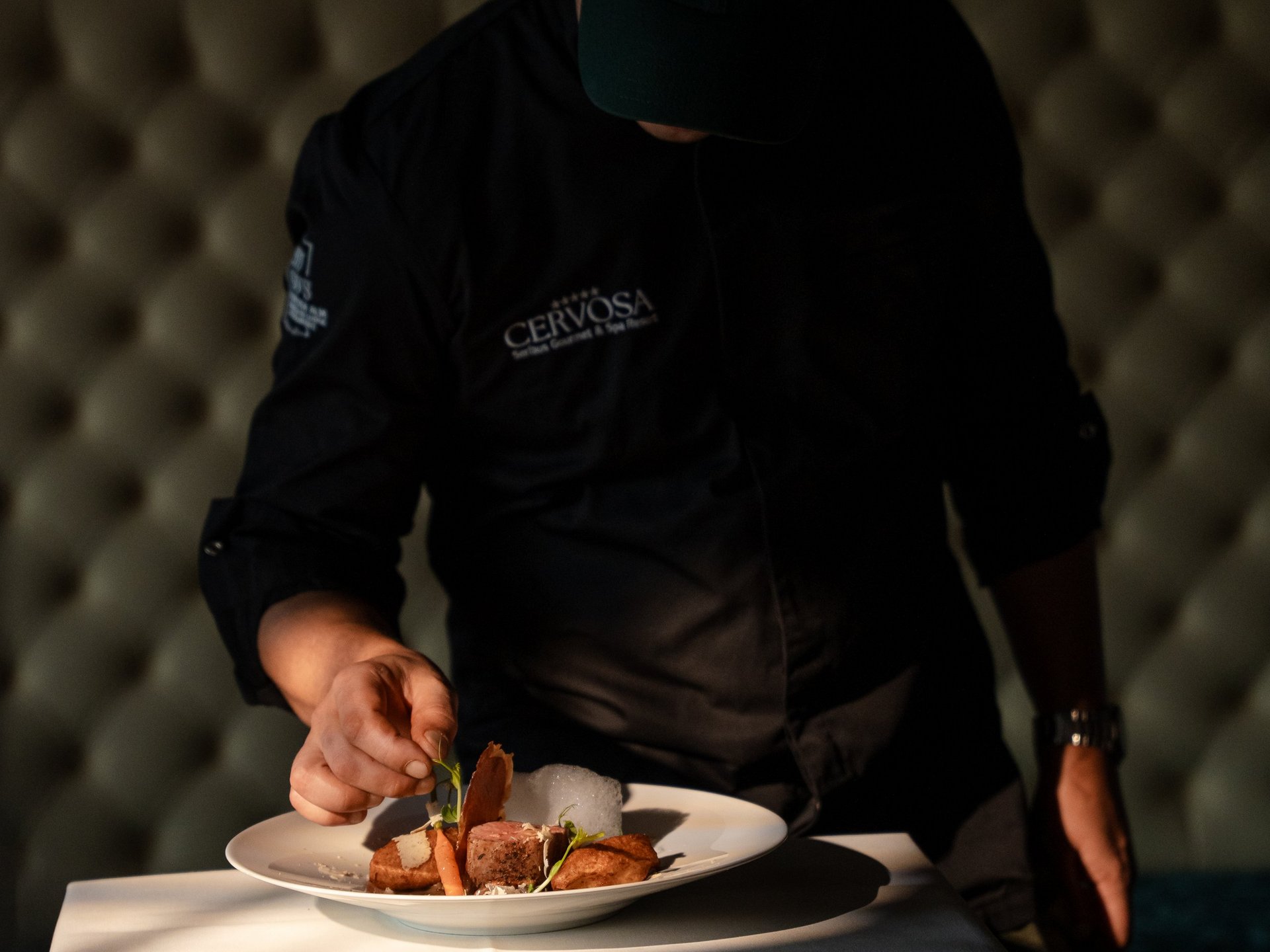Chef wearing baseball cap artfully plating a dish on a white table with tablecloth