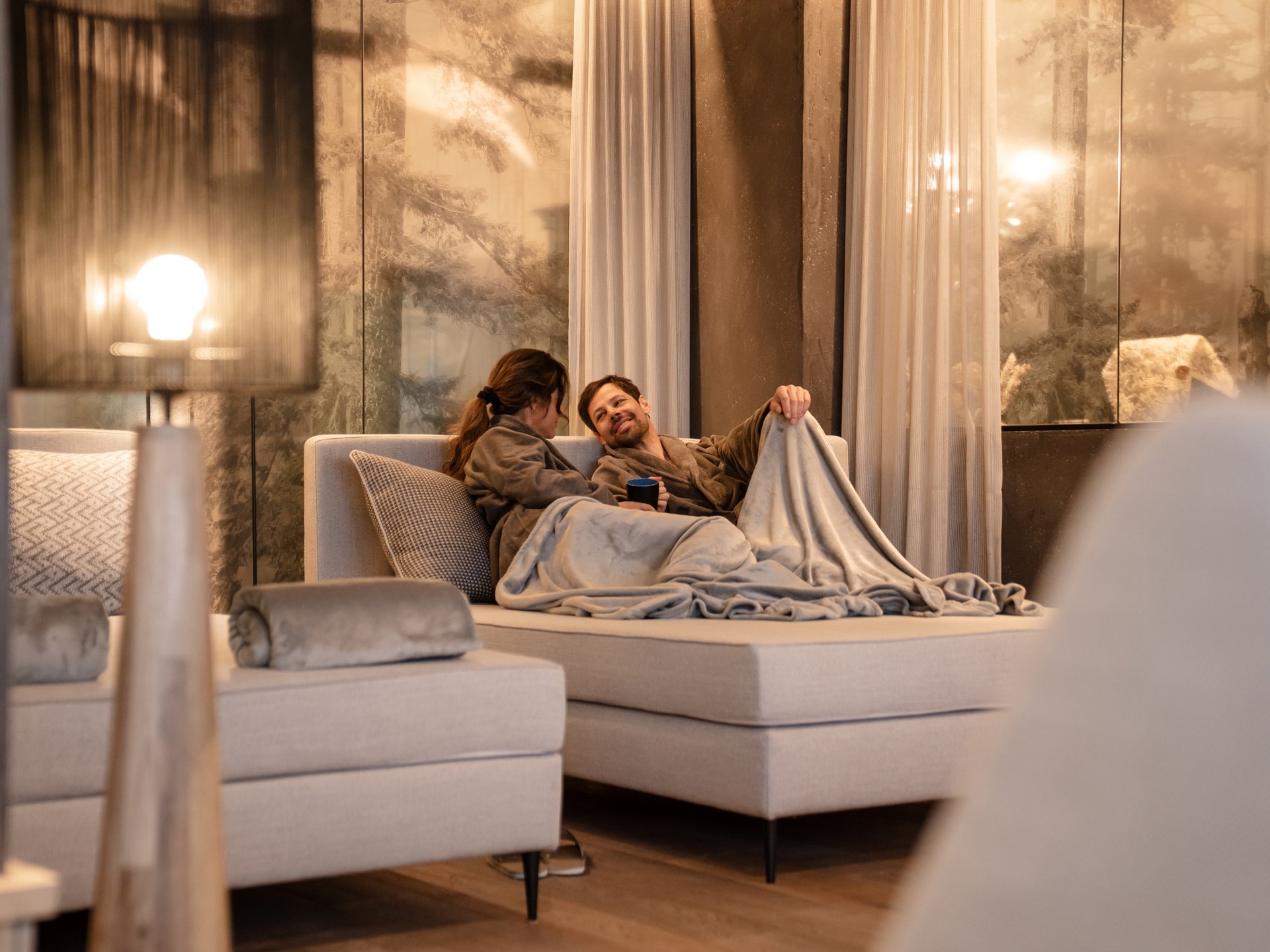 Wohlfühlurlaub im 5-Sterne-Hotel in Serfaus Paar in Bademänteln kuschelt sich auf Sofa in gemütlichem Raum mit warmem Licht