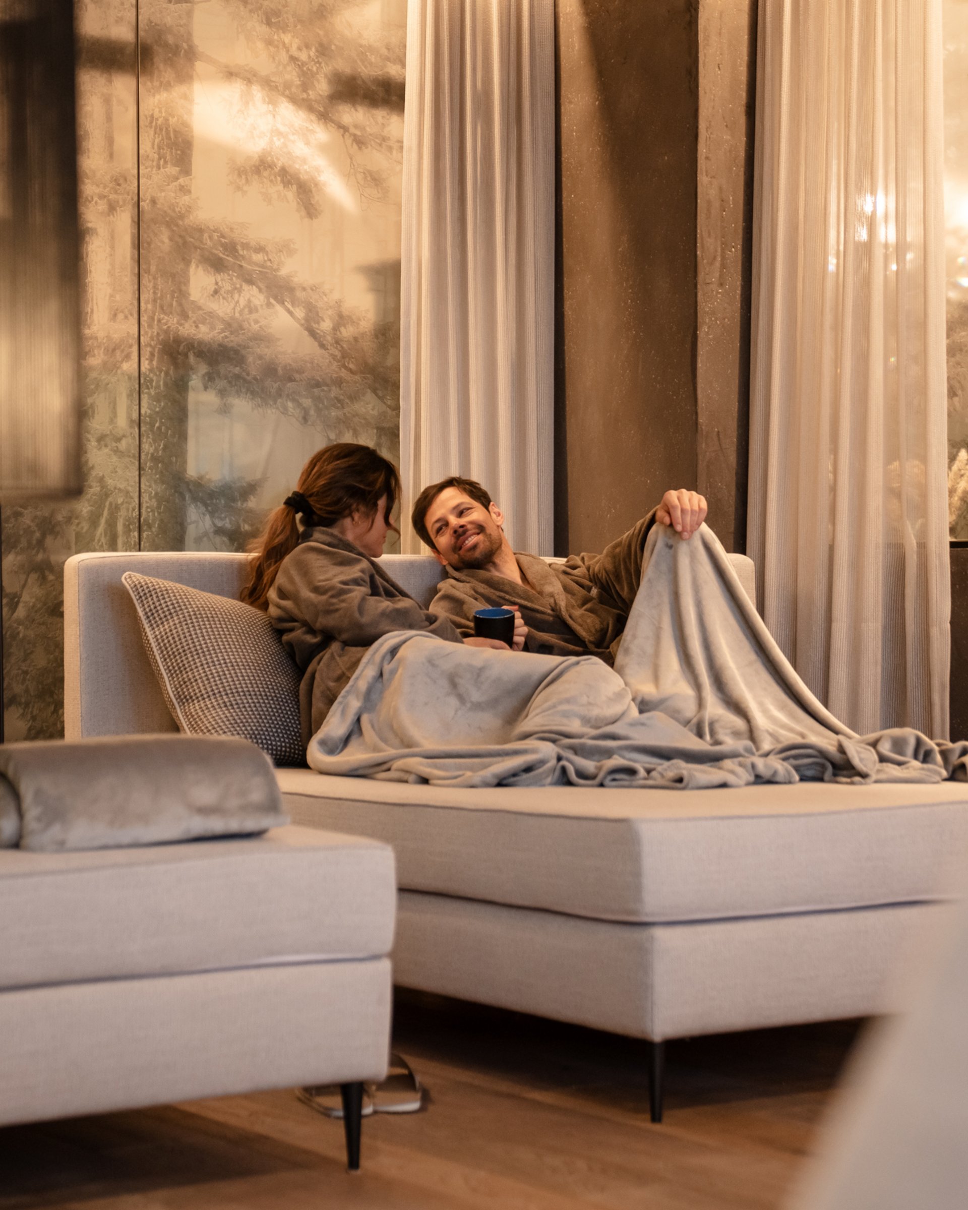 Paar in Bademänteln kuschelt sich auf Sofa in gemütlichem Raum mit warmem Licht