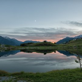 Bergsee spiegelt Hügel und Mond in ruhiger Abenddämmerung