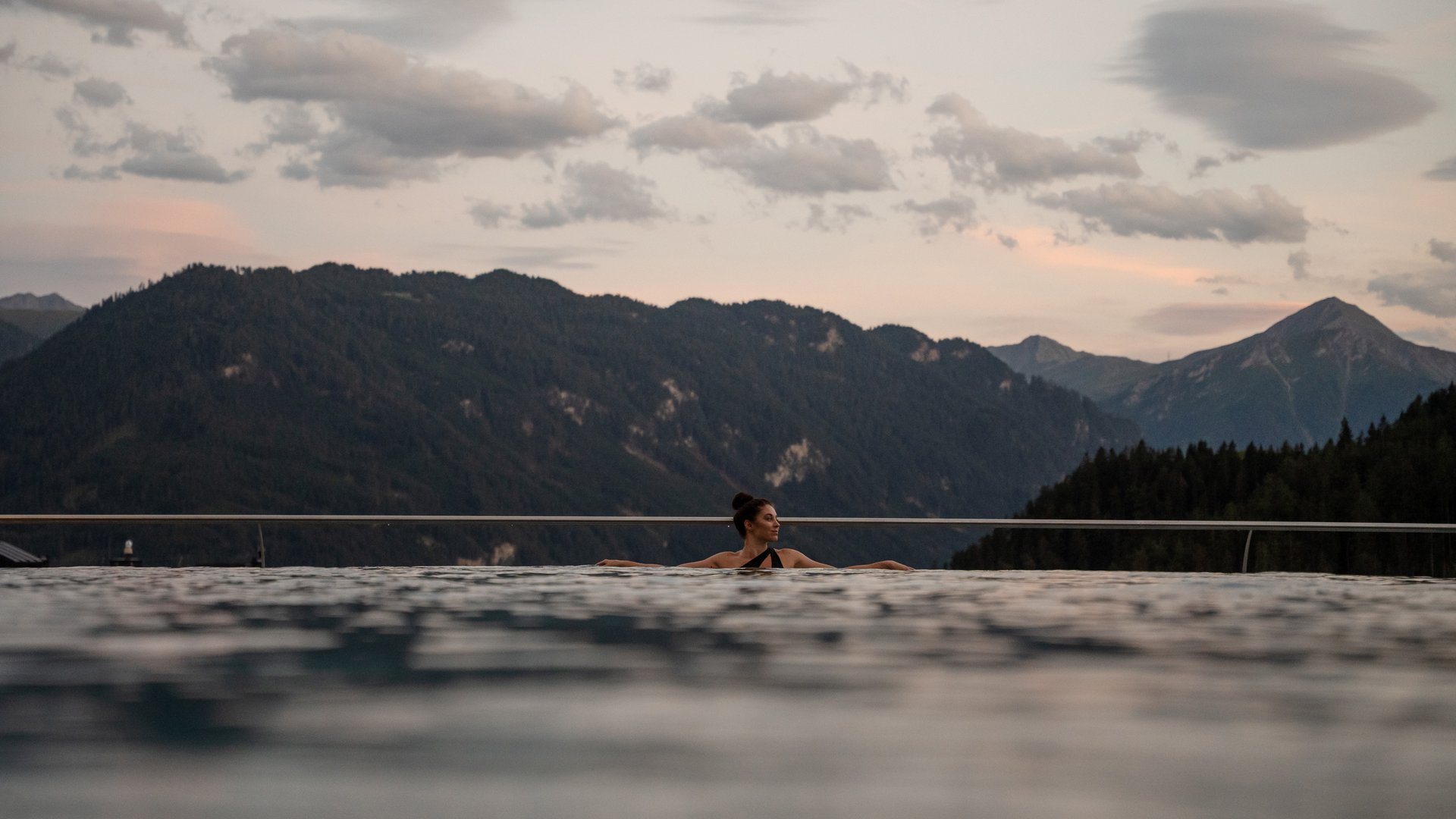 Woman relaxing in infinity pool with forested mountains in the background at sunset