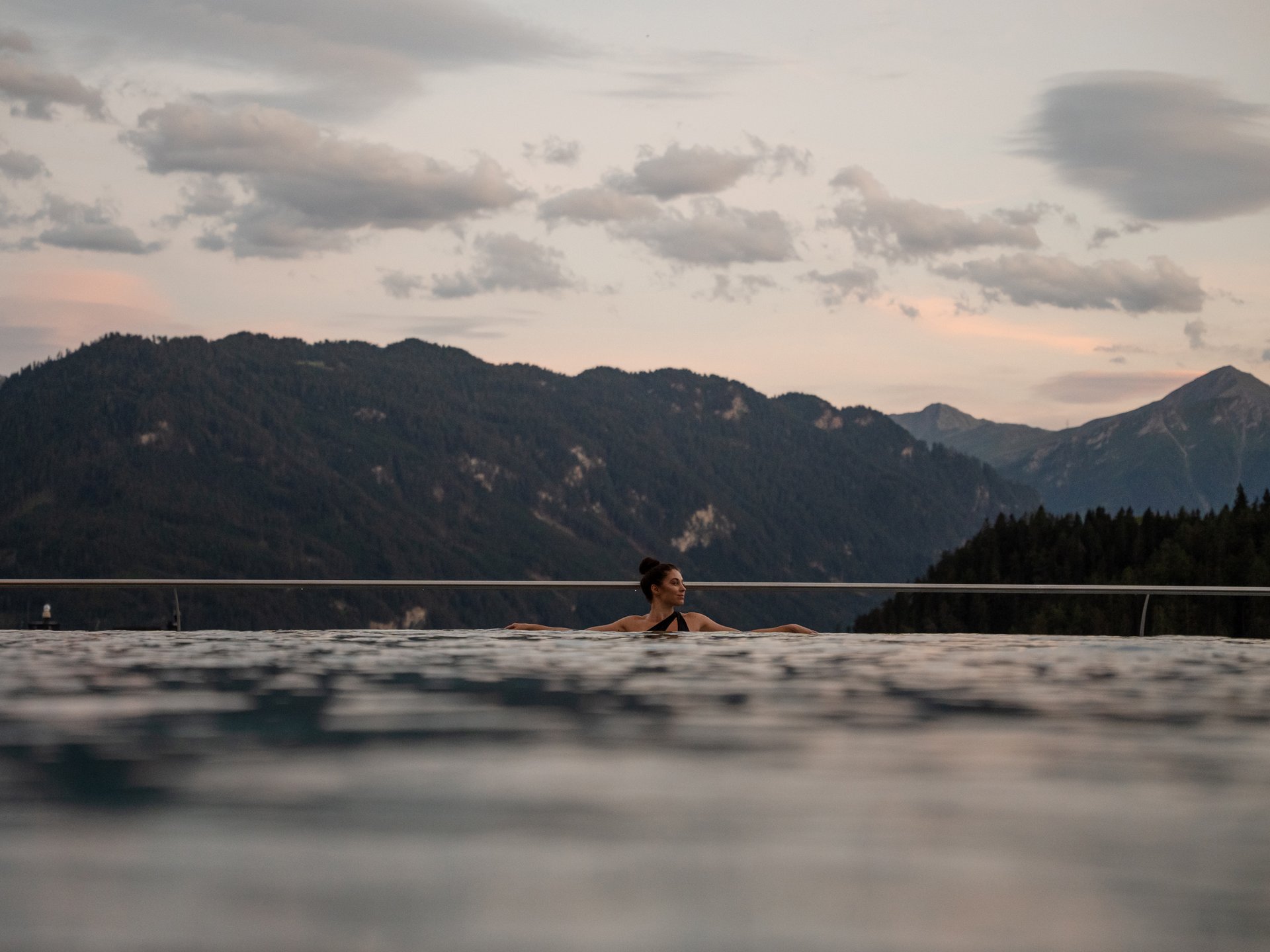 Bathing bliss at our hotel with pool in Serfaus Woman relaxing in infinity pool with forested mountains in the background at sunset