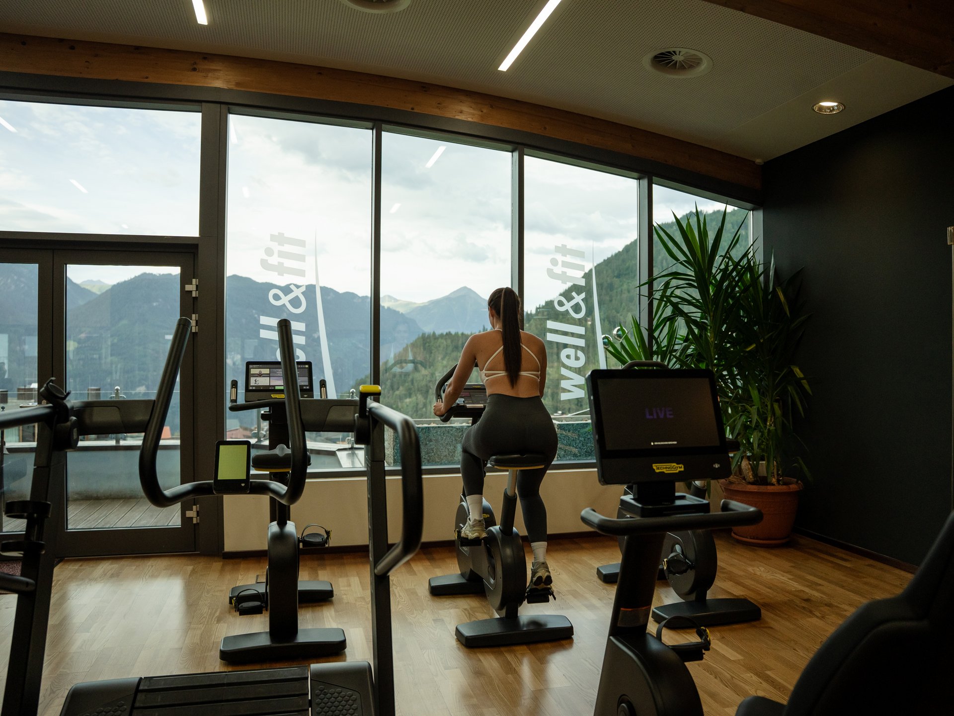 Bewegung im Sporthotel mit Fitnessstudio Frau trainiert auf Heimtrainer vor Panoramafenster mit Bergblick im Fitnessraum