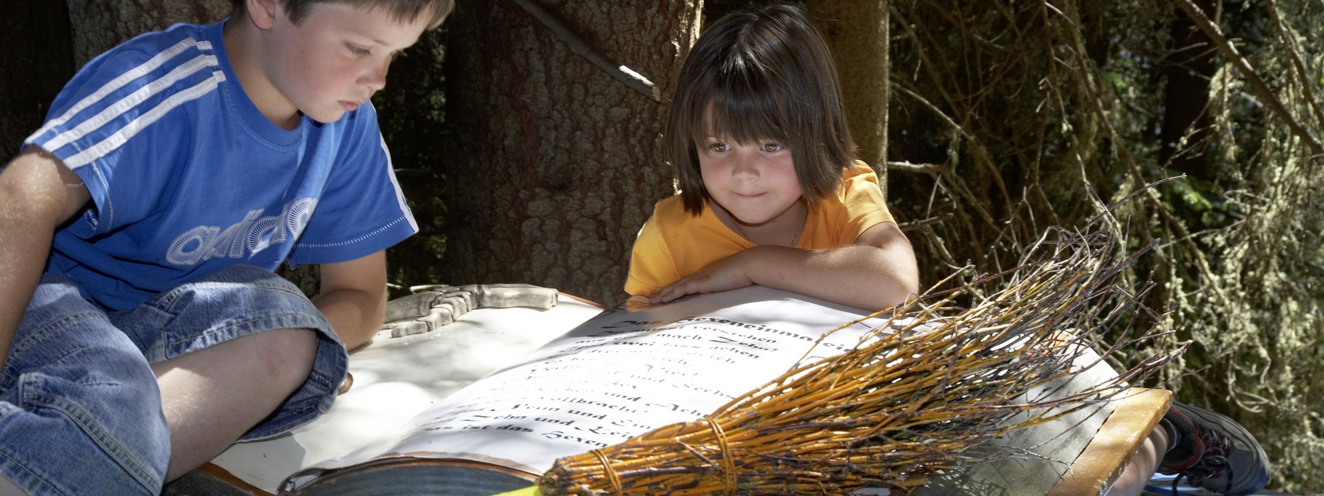 Zwei Kinder lesen ein großes Buch im Wald neben einem Besen