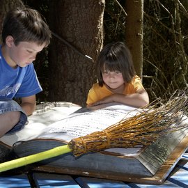 Zwei Kinder lesen ein großes Buch im Wald neben einem Besen