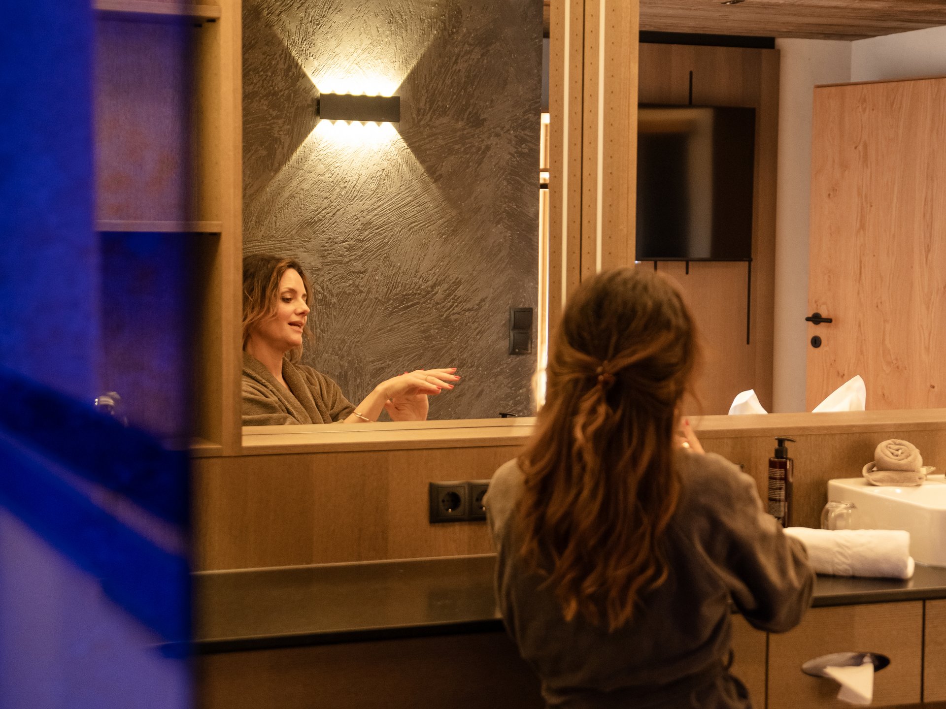 Frau im Bademantel sitzt vor Spiegel in modernem Badezimmer.