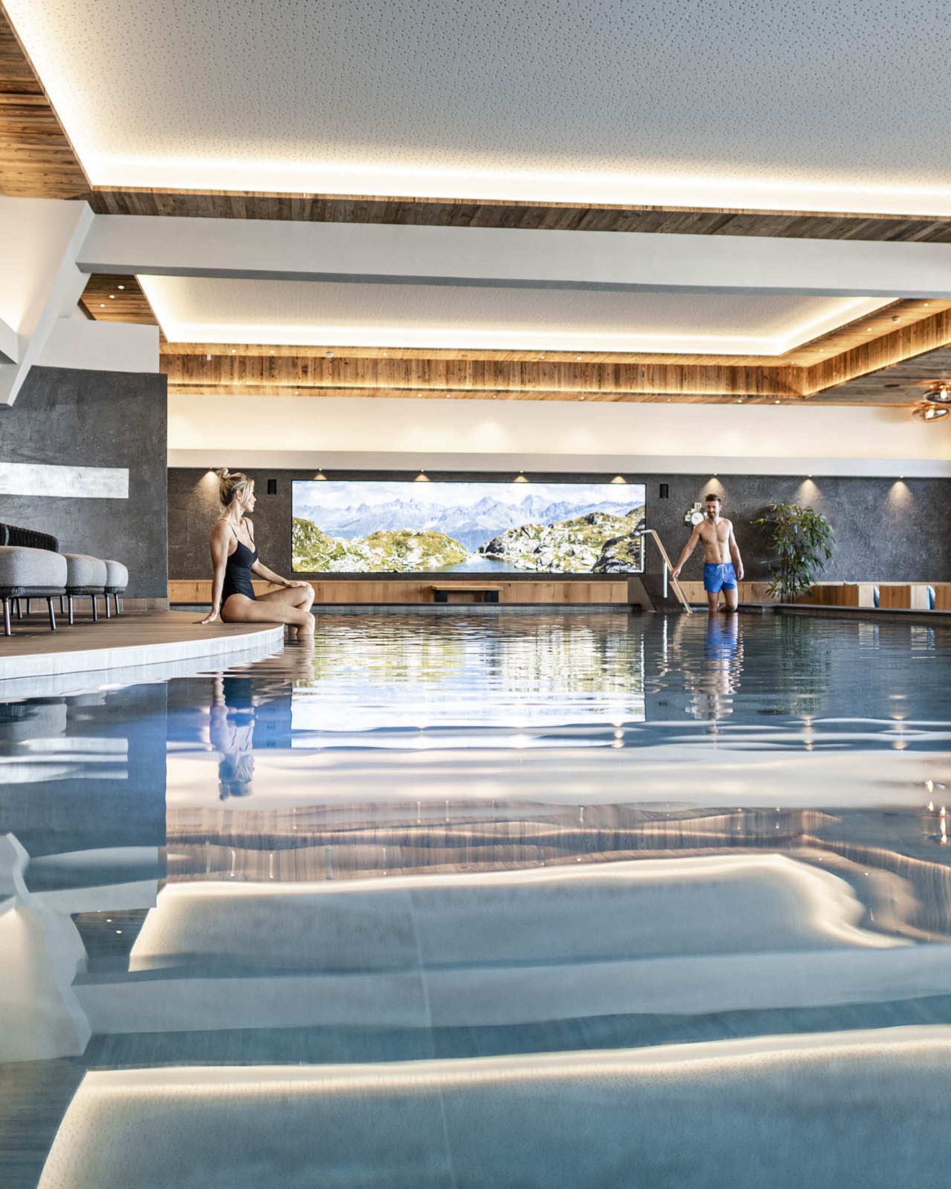 Innenraum mit Schwimmbad, Frau sitzt am Beckenrand, Mann steigt ins Wasser