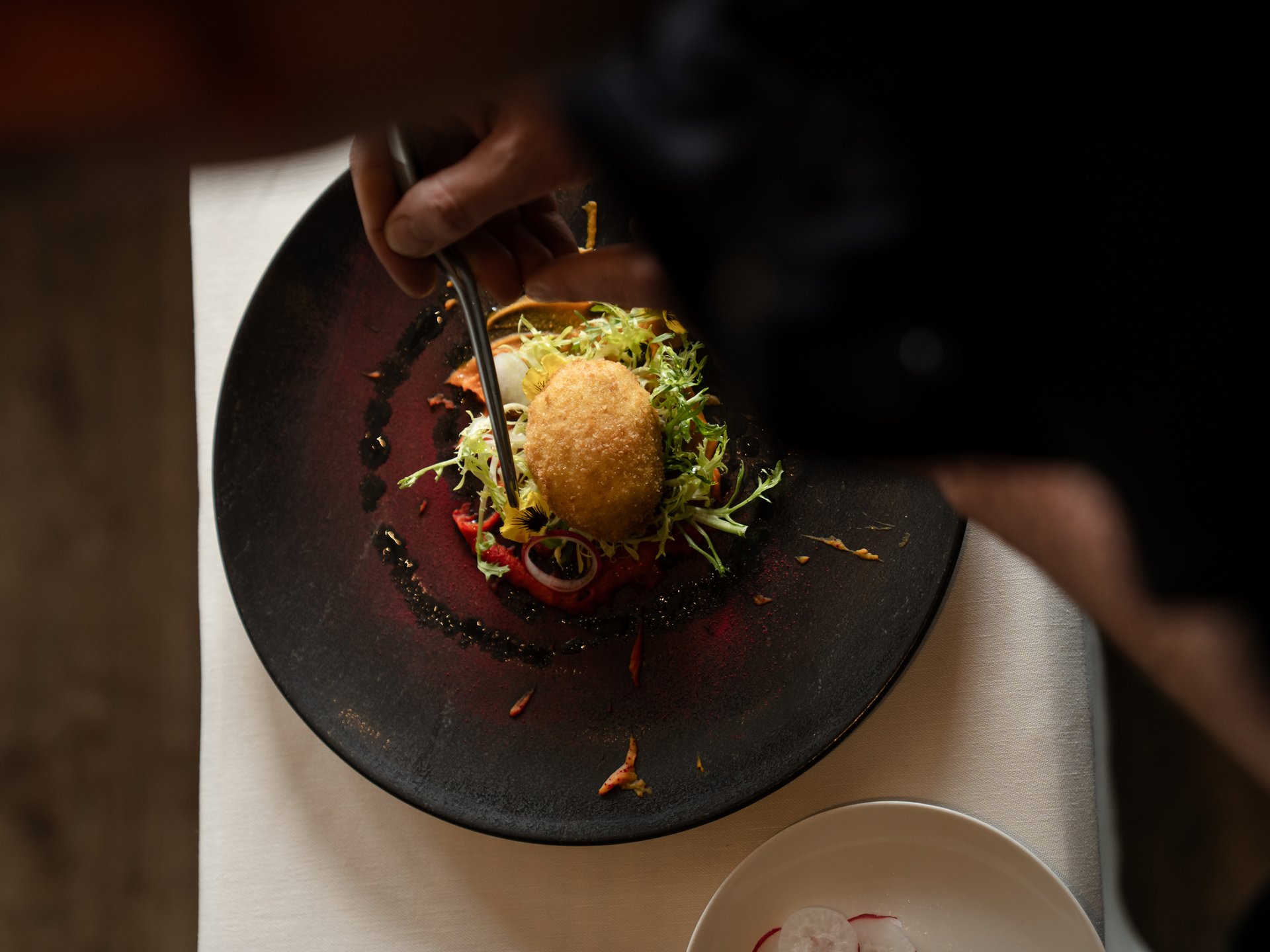 Der Gipfel des Geschmacks im Gourmethotel in Tirol Hand verwendet Besteck, um auf einem schwarzen Teller Salat mit einem panierten Gericht zu essen