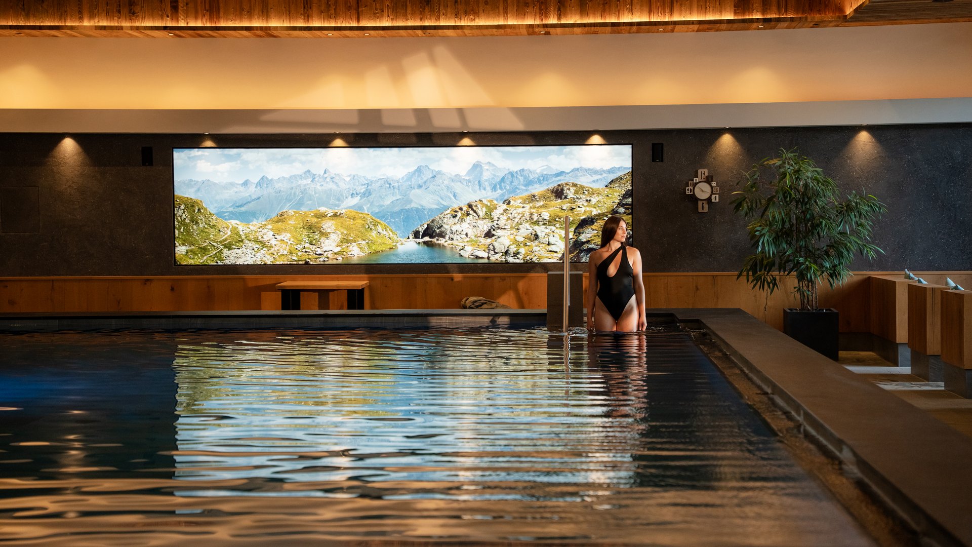 Woman in black swimsuit standing in indoor pool with mountain view window