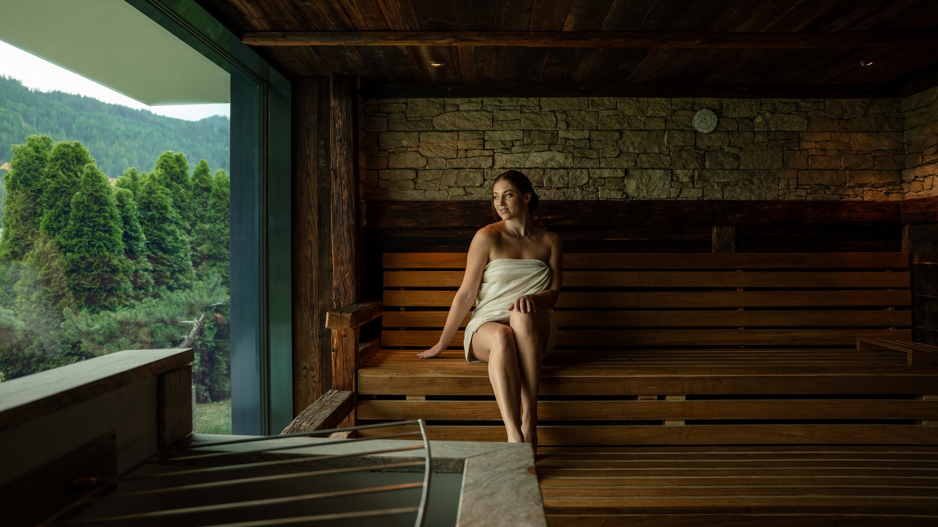 Woman wrapped in towel relaxing in sauna with view of green trees