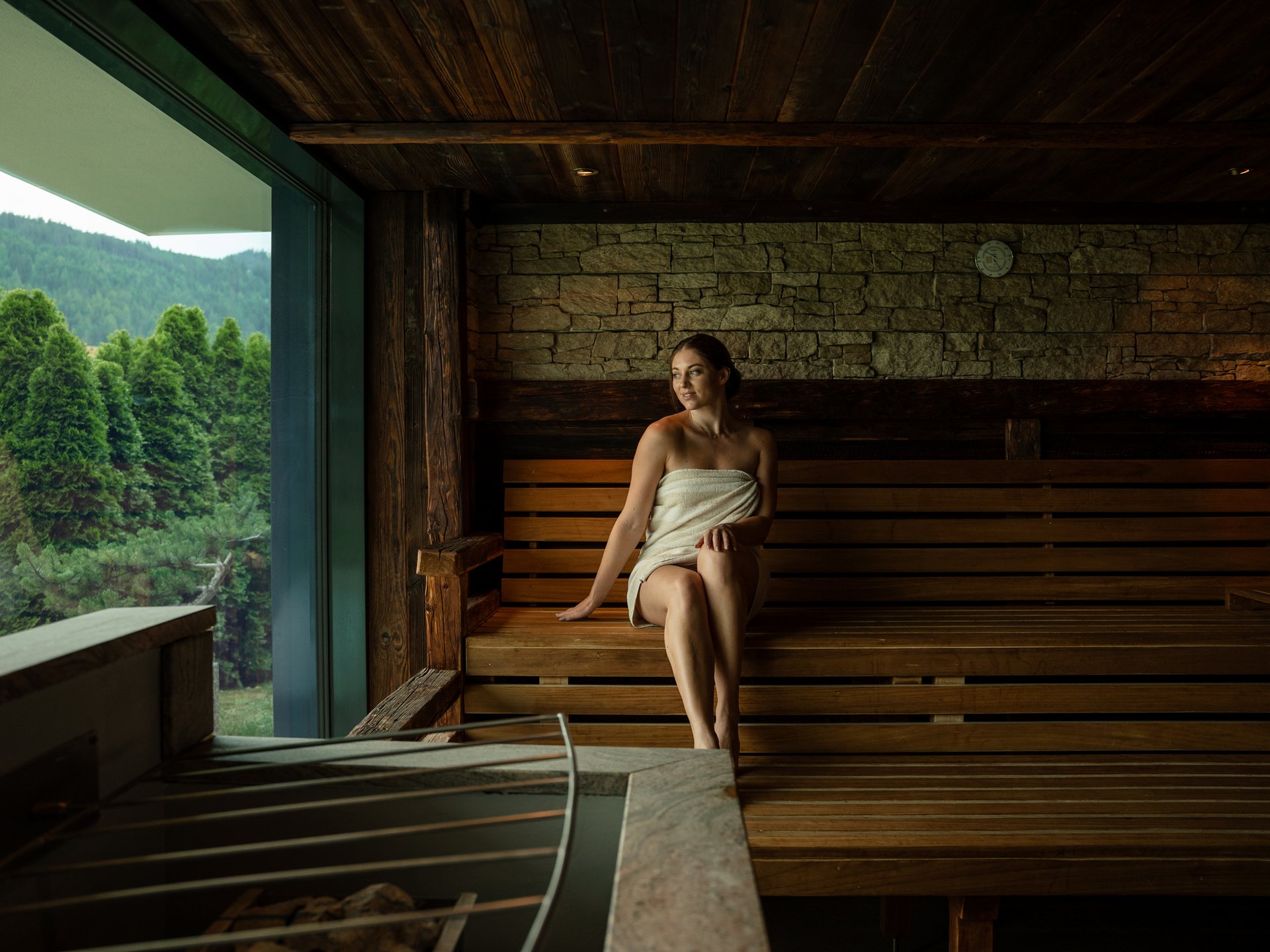 Feel-good holiday at our 5-star hotel in Serfaus Woman wrapped in towel relaxing in sauna with view of green trees