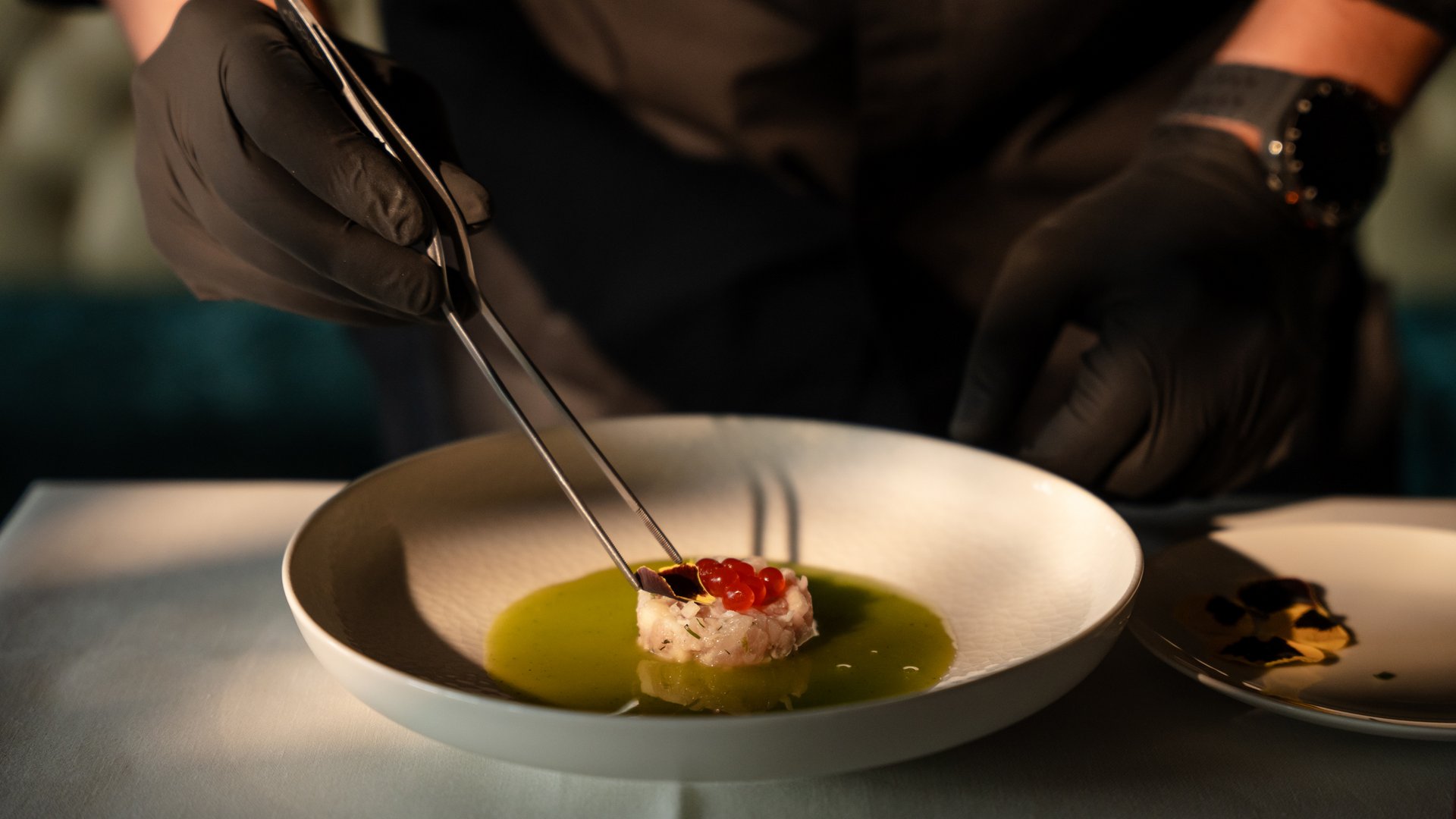 Chef wearing black gloves decorates fine dish with tweezers