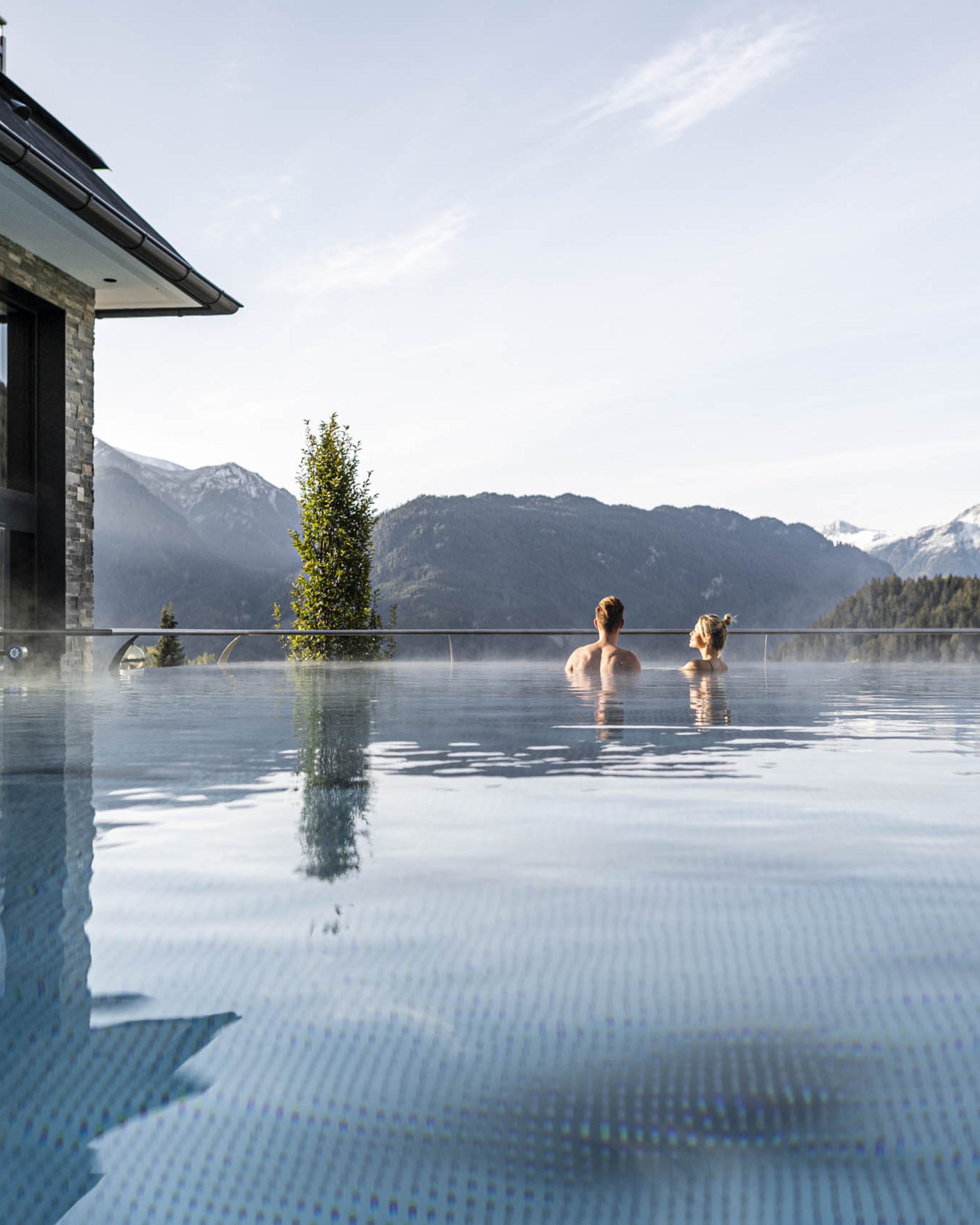 Two people in an outdoor pool with mountain views and misty sky