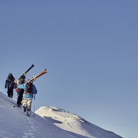 Drei Skifahrer wandern einen schneebedeckten Hang bei klarem Himmel hinauf