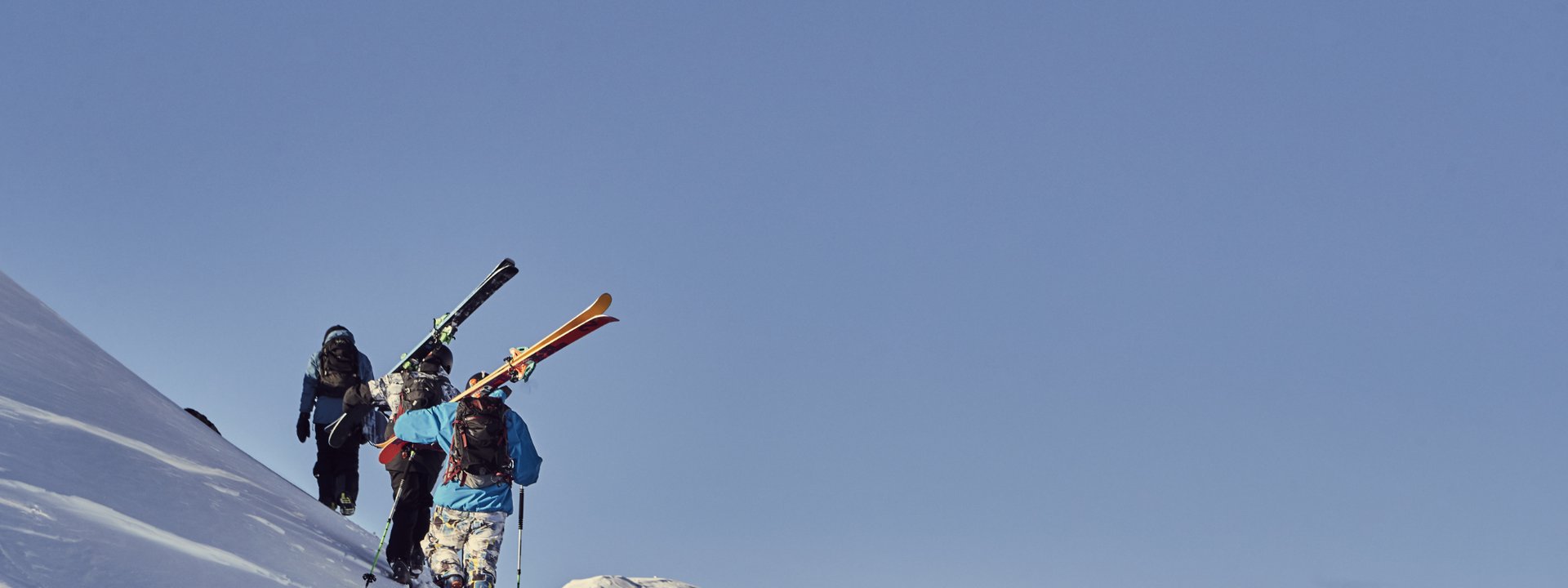 Drei Skifahrer wandern einen schneebedeckten Hang bei klarem Himmel hinauf